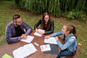 Students are sitting outside at a wooden table, on which books and study documents lie.