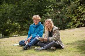 A blond student with a blue jacket and a blonde student with a brown jacket are sitting in a meadow.